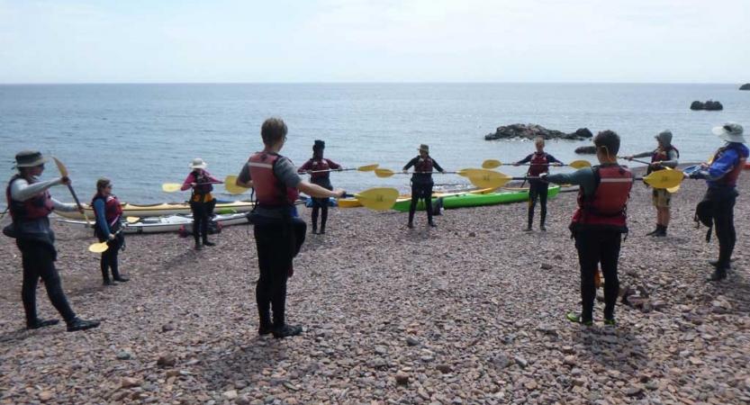 A group of people stand in a circle holding paddles in an apparent kayaking exercise. Several colorful kayaks rest on the shore with a vast lake in the background. 